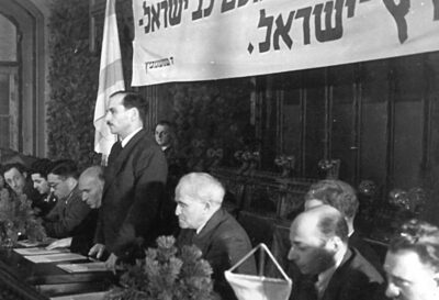 Dr. Zalman Grinberg (stehend), Überlebender aus dem KZ Kaufering, begrüßt die Delegierten und den Ehrengast David Ben Gurion (4. v. r.) zur Sitzung des Zentralkomitees der befreiten Juden im Münchner Rathaus. Foto: hagalil.com
